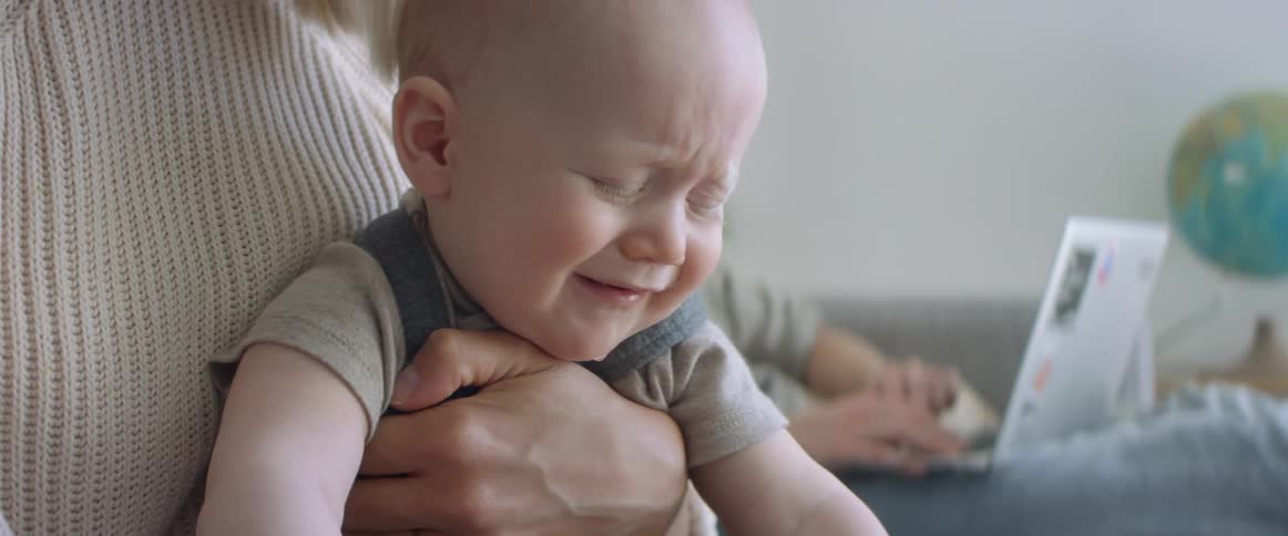 A woman holds her crying baby as her husband sits with his laptop on his knee