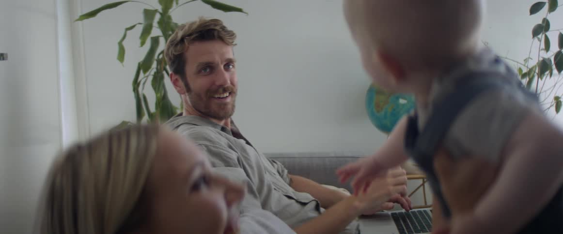 A mother sitting on a couch lifts her baby son up in the air and his dad reaches over and tickles him