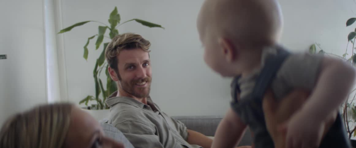 A mother holds up a baby in the air with the father sitting across from them on the couch