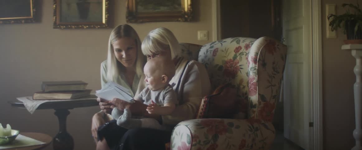 Granddaughter sitting on grandmothers lap while the daughter sits beside her as the grandmother looks through photos and they all smile and laugh together