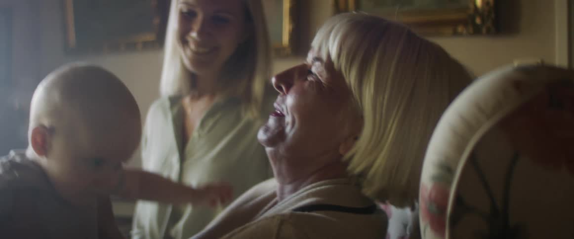 A grandmother bounces a baby on her lap as his mom watches them and smiles