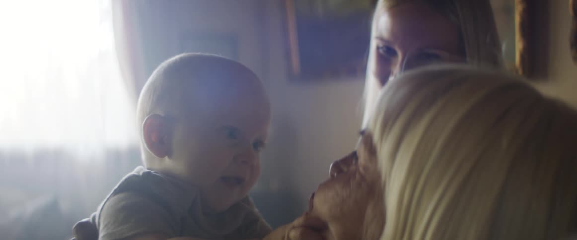 A grandmother bounces her grandson on her knee and talks to him and laughs as his mom watches and smiles