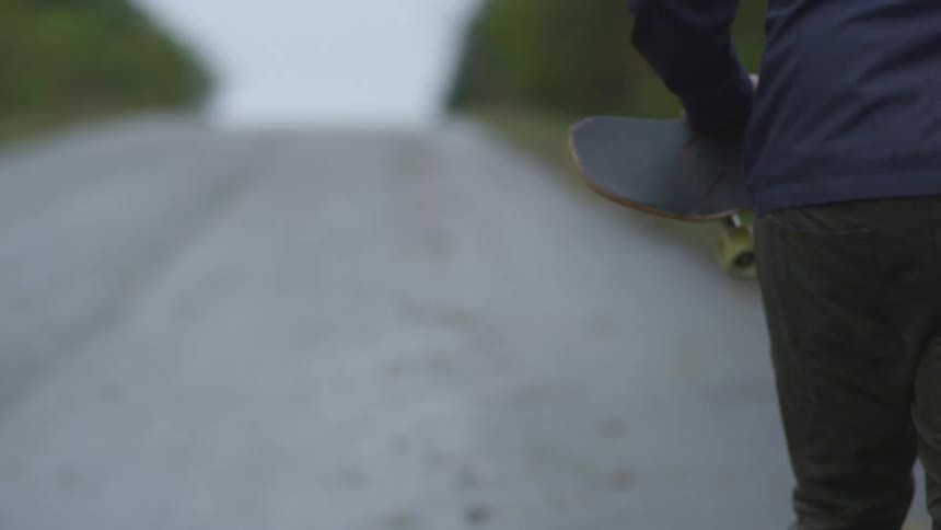 Person walks along empty road through trees carrying skateboard