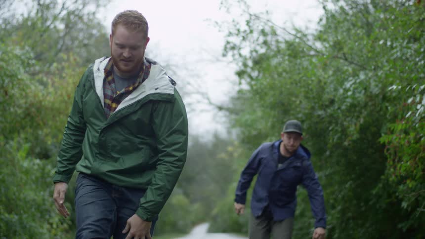 Two men ride skateboards on a path surrounded by trees on an overcast day