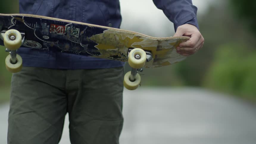 A man walking down a road turns around and hold his skateboard in both hands