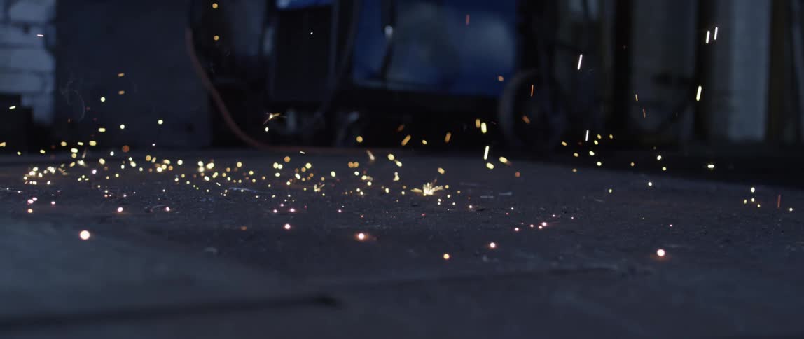 Sparks fall onto dark floor in shadowy building with machinery near wall