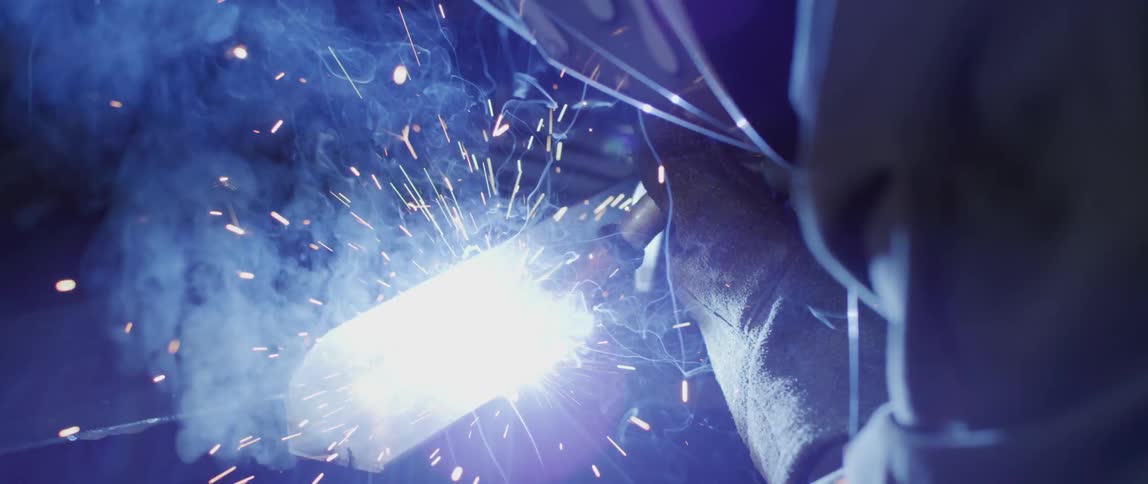 A person with a welding helmet and gloves welding a piece of metal with a torch