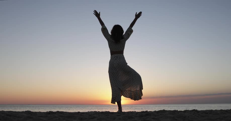 Woman moves through a yoga pose while standing on beach facing sun sitting on horizon and turning sky red over ocean