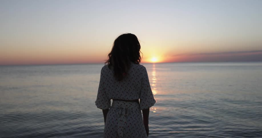 Woman wearing dress stands looking across ocean toward sun on horizon under red sky and is silhouetted by sunshine