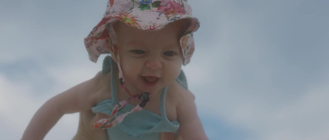A person holding up a baby wearing a hat with the sky and clouds in the background