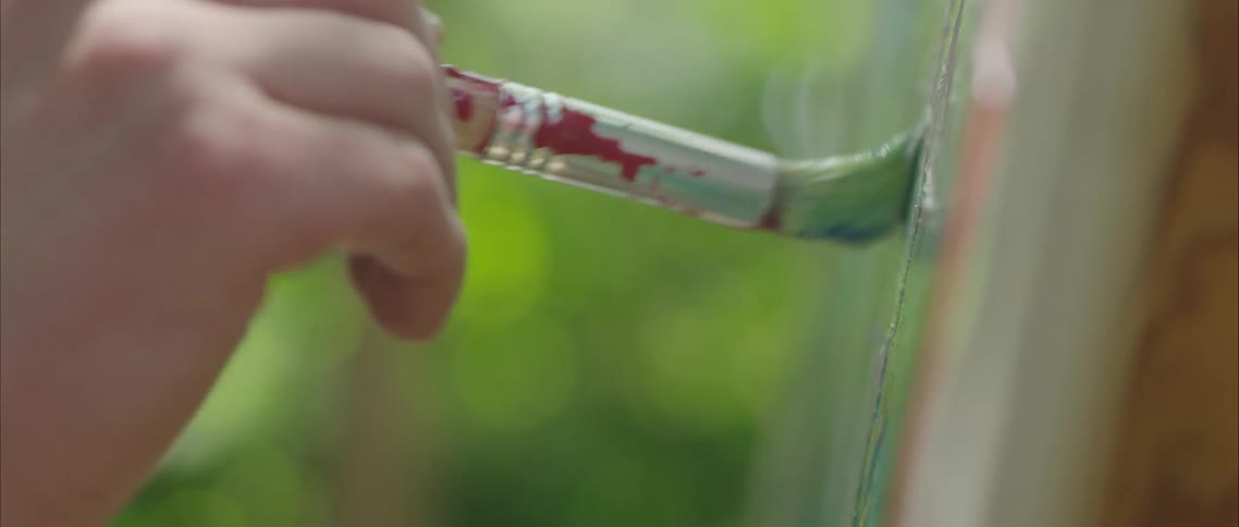 A person uses a stained paintbrush to paint on a canvas outdoors during the day