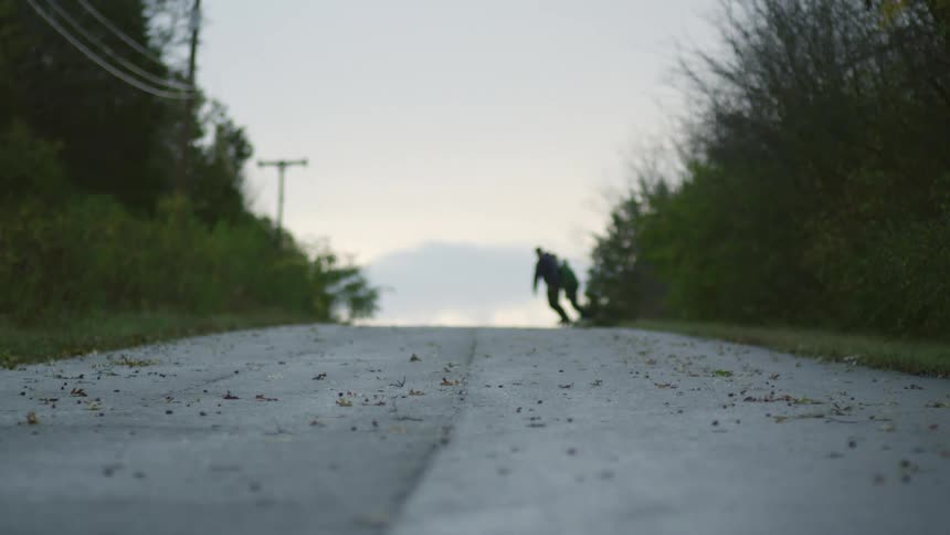 A pair of skateboarders zigzag across a road as they skateboard on a cloudy evening