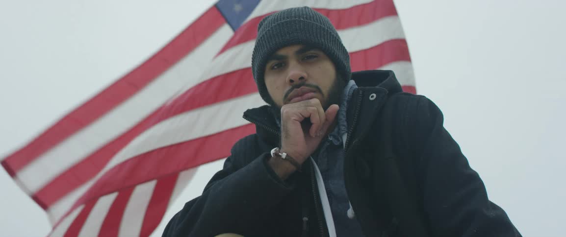 A man in a knit hat and coat poses with his chin on his hand in front of a large american flag