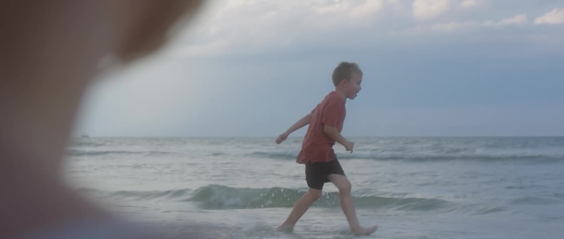 A boy running in ocean waves on a beach while his mother watches on