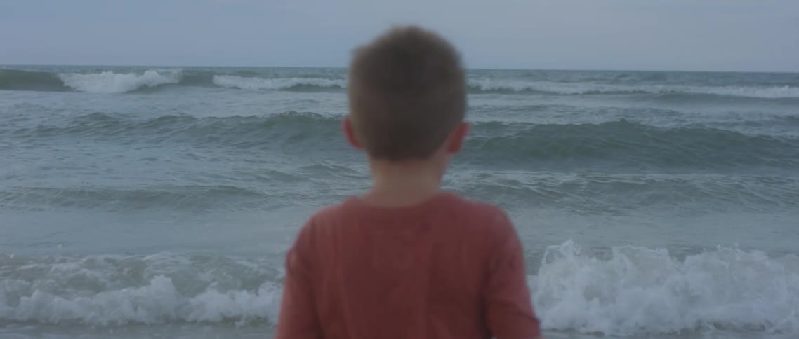 A boy standing on the beach with waves the sky and clouds in the background