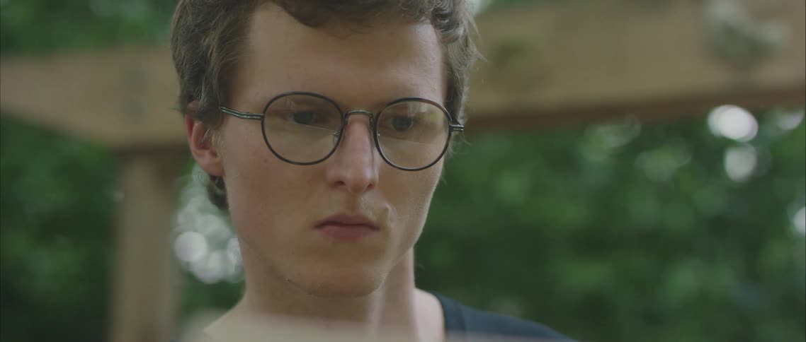 A man wearing round eyeglasses uses his laptop on a porch outside