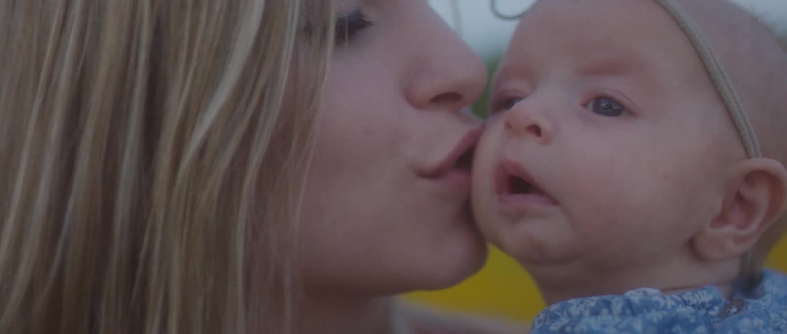 A mother holds her baby girl outside and kisses her cheek and smiles