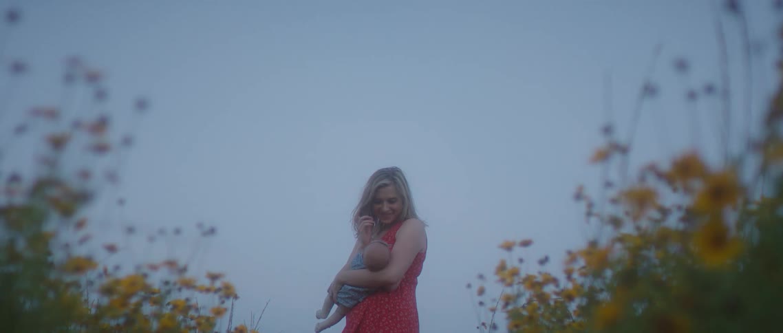 A woman rocks a baby in her arms while standing between rows of yellow flowers in a field under a clear blue sky