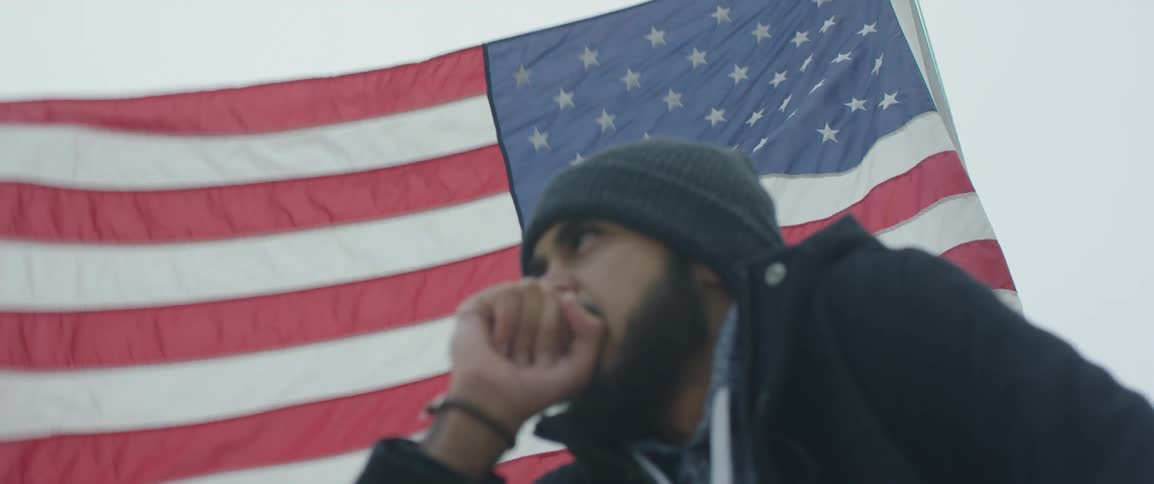 A man wearing a coat and knit hat sits in front of a large american flag with his face resting against his fist