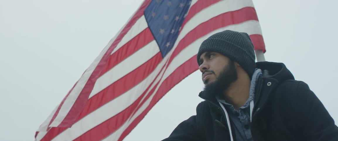Bearded man wearing a beanie hat stands besides a waving american flag
