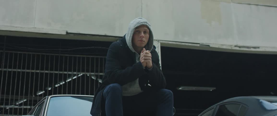 A man in a hooded sweater and coat sits on top of a vintage car parked outside a garage