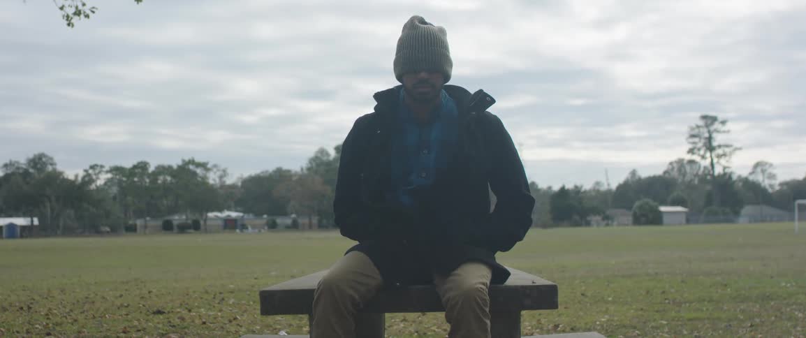 A man in a coat and knit cap sitting on a park bench on a cloudy day