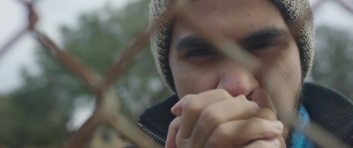 Man stands outdoors looking through chain link fence clasping hands near mouth