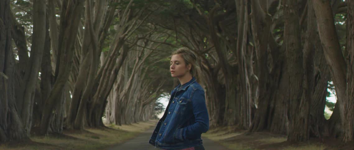 A woman in a denim jacket standing beneath mature trees with her hands in her pockets