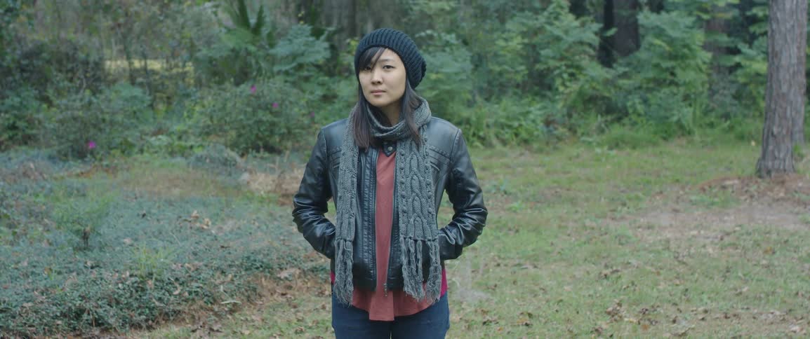 A woman stands in a forest wearing a jacket scarf and knit hat