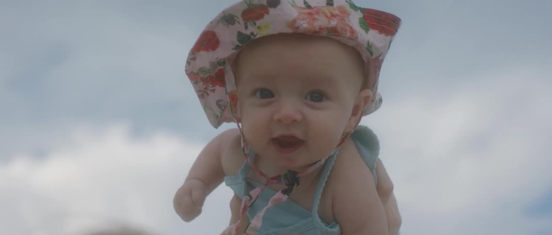 A person holds a small infant girl up in the air and she is wearing a sunhat and bathing suit