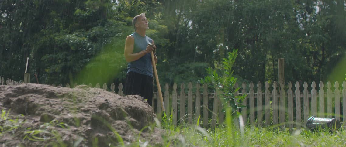 An elderly man rests against a shovel driven into the dirt in a fenced yard on a rainy day