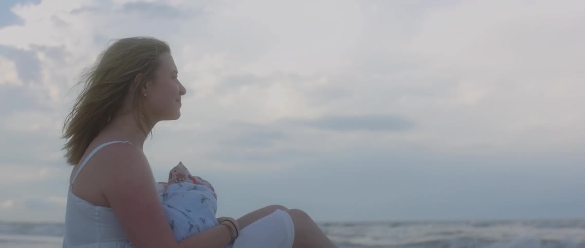 A woman smiles as she cradles her baby while sitting on the beach next to the ocean
