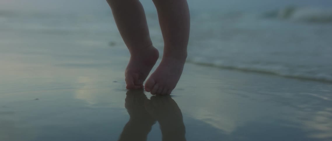 A baby being lifted in the air is slowly lowered to sands of a beach until an ocean wave rolls of its feet