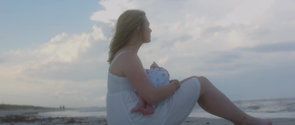 A woman sitting on a beach holding a baby with the ocean sky and clouds in the background