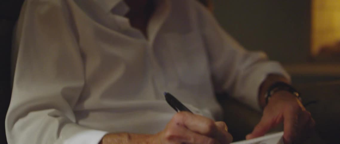 A man writes in a notebook while sitting in a brown armchair by the fireplace