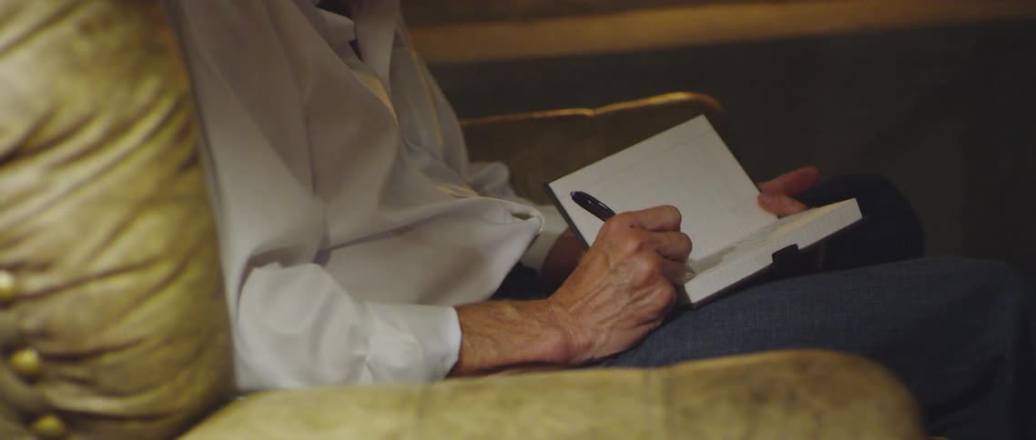 An elderly man writes in a journal while sitting on a cushioned chair in front of a fire place in a house
