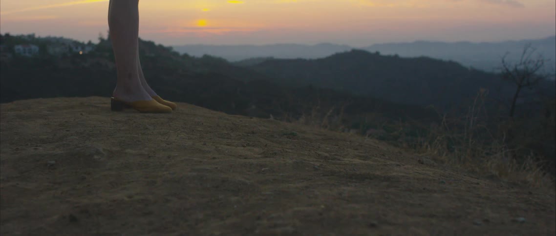 The legs of a woman standing atop the barren soil on the top of a hill in a hilly countryside at sunset