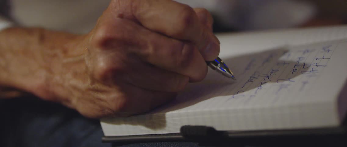 An elderly person writes in a leather bound notebook with a blue pen