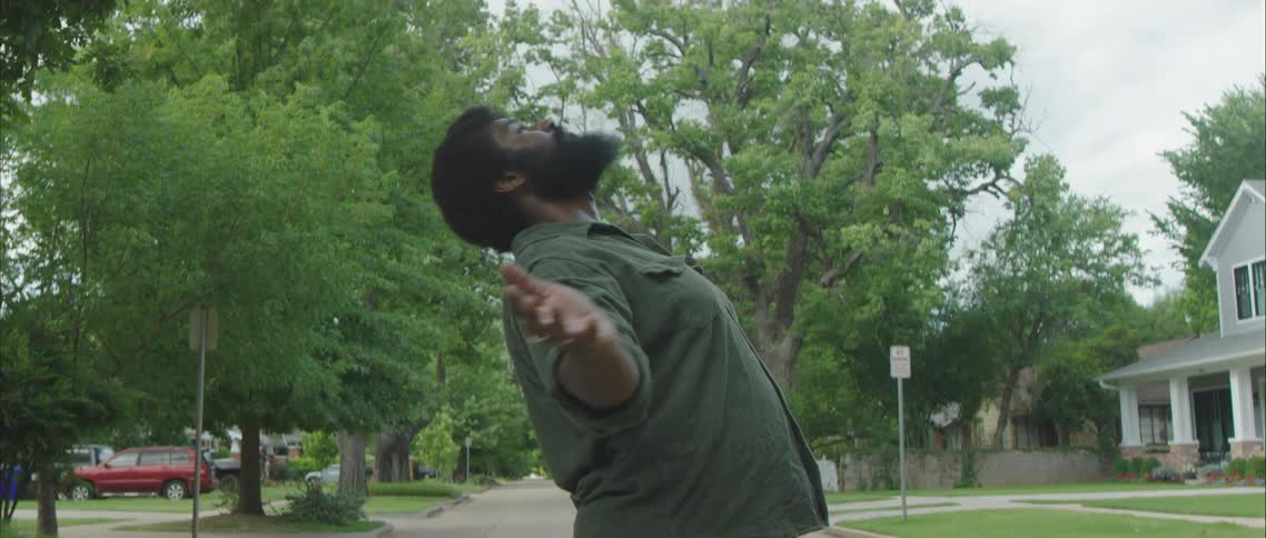 A man performs a dramatic dance move while standing in the center of a road in a residential neighborhood during the day