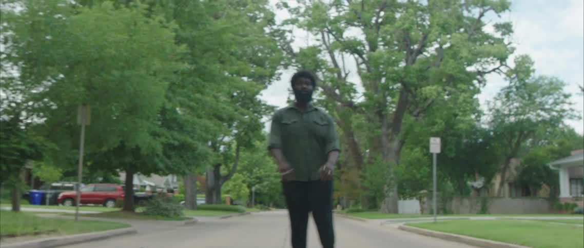A large black man dances in the road of a quiet neighborhood on a spring day