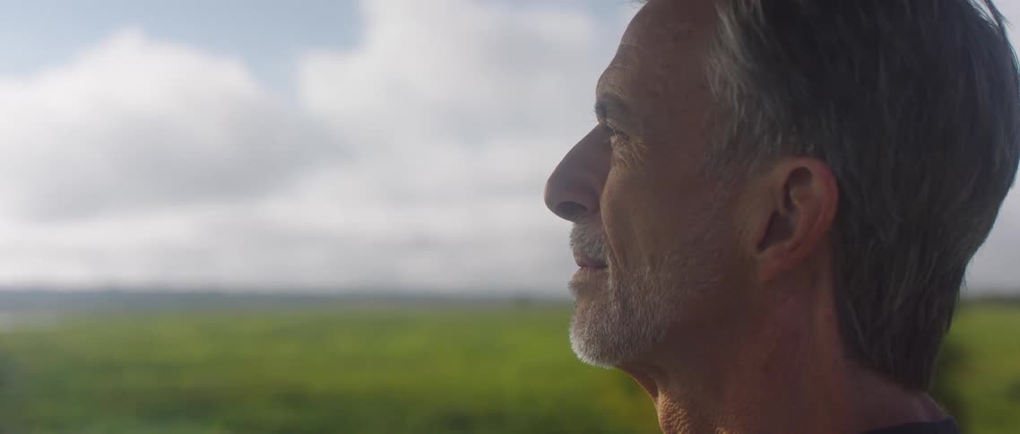 A man looking off into the distance with trees a grassy field the sky and clouds in the background