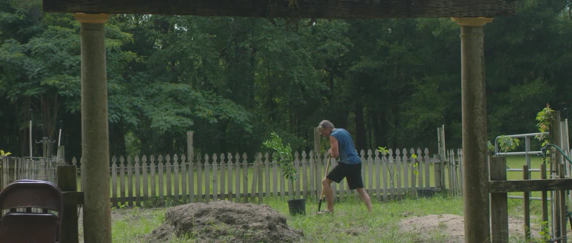 A man uses a spade dig in a yard surrounded by a wooden fence during the day