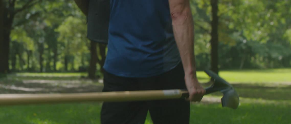 Man carries a potted plant out to a grassy area outdoors while carrying a shovel