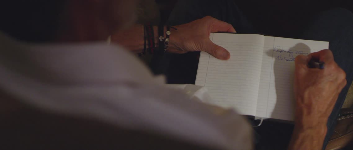 An elderly man writing in a notebook on his lap