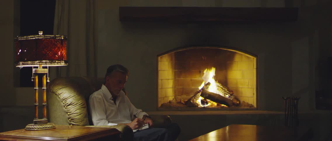 Man wearing a white shirt sitting on a chair near the fireplace in a mansion at night