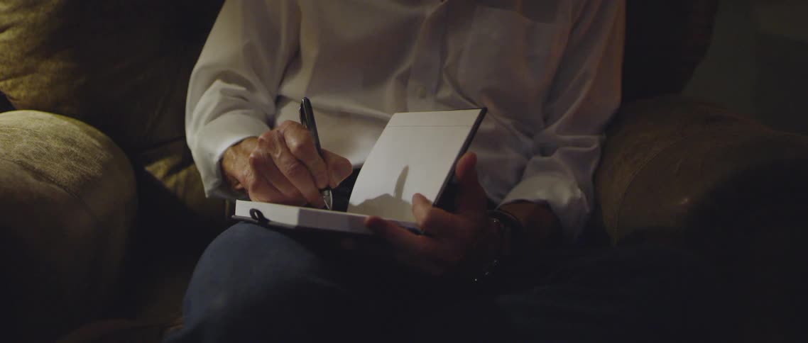 An elderly man writes in a leather bound journal with a pen while sitting on a chair in a house