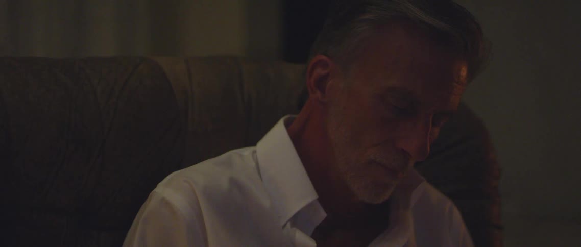 Flames from a fireplace flicker across the forehead of a man sitting in a large armchair