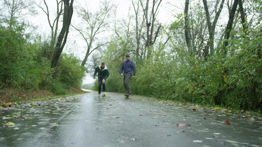 Two men skateboard on a wet paved path in overcast weather