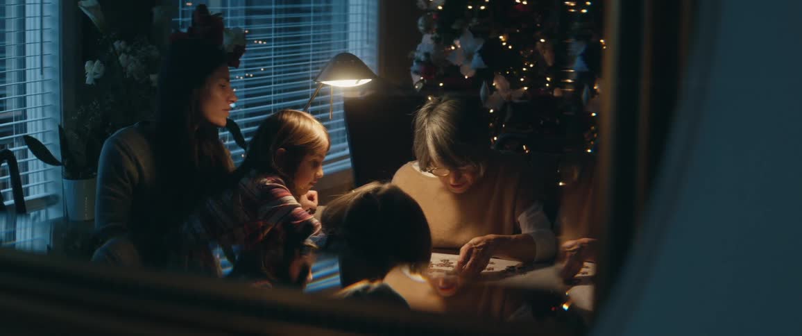 Mom daughters and grandmother sit at a table putting a puzzle together in front of a christmas tree