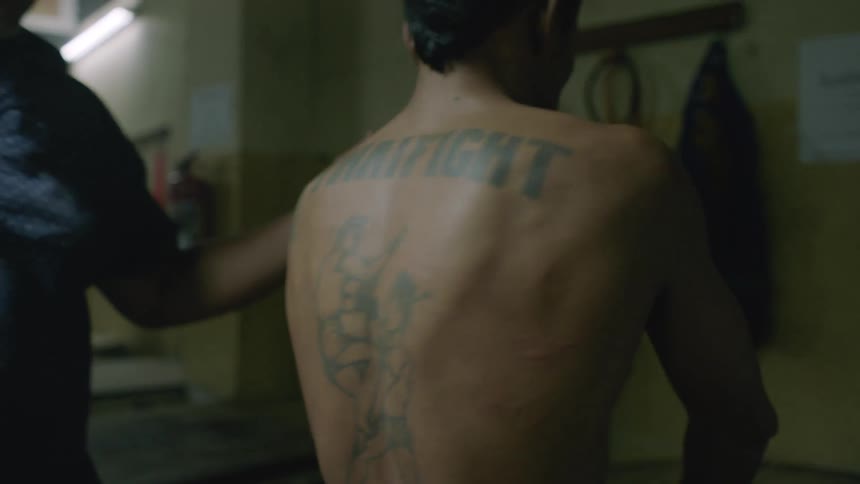 A fighter with a large back tattoo lies back on a table as his coach places a towel over his face